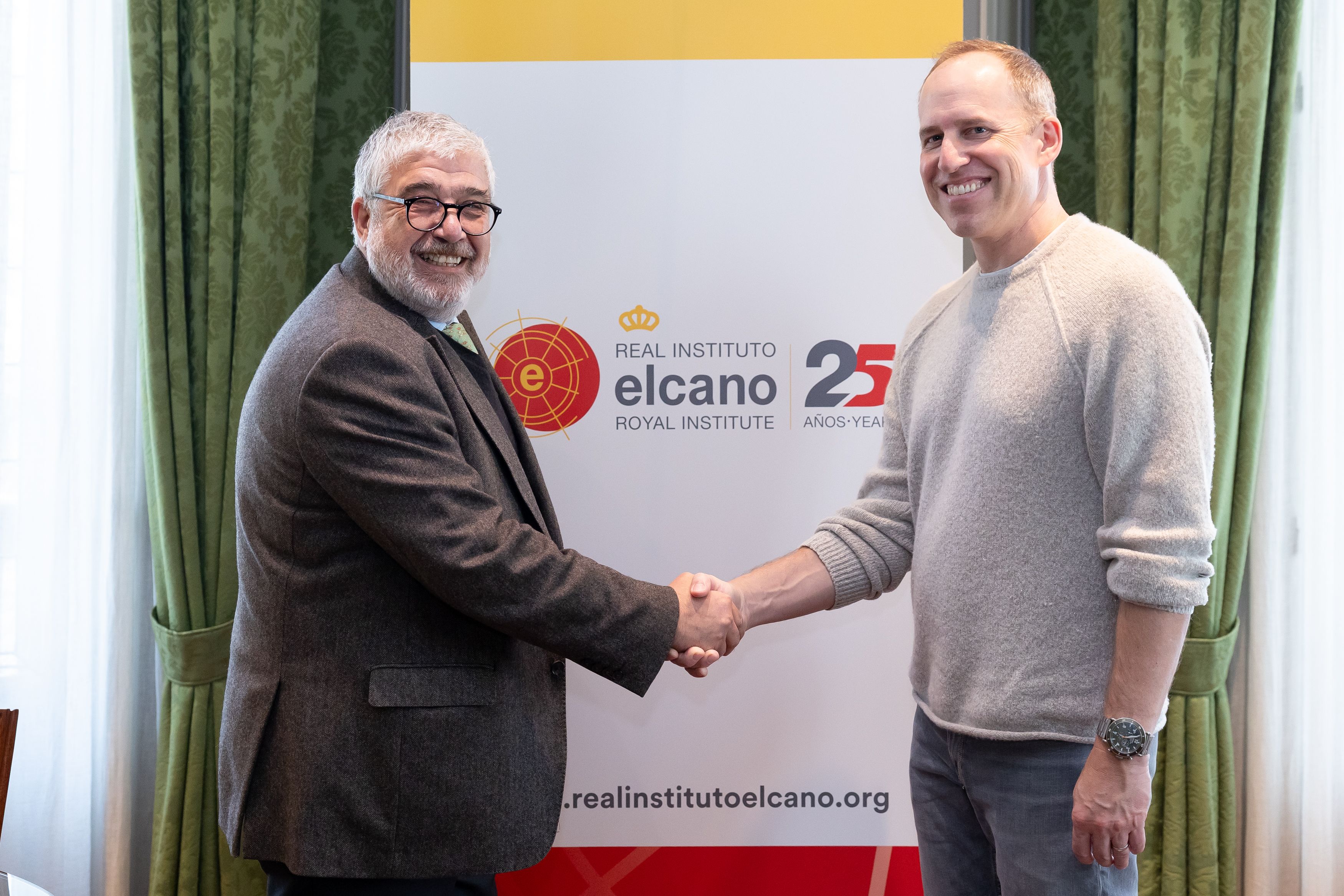Two smiling men shaking hands in front of an Elcano Royal Institute banner