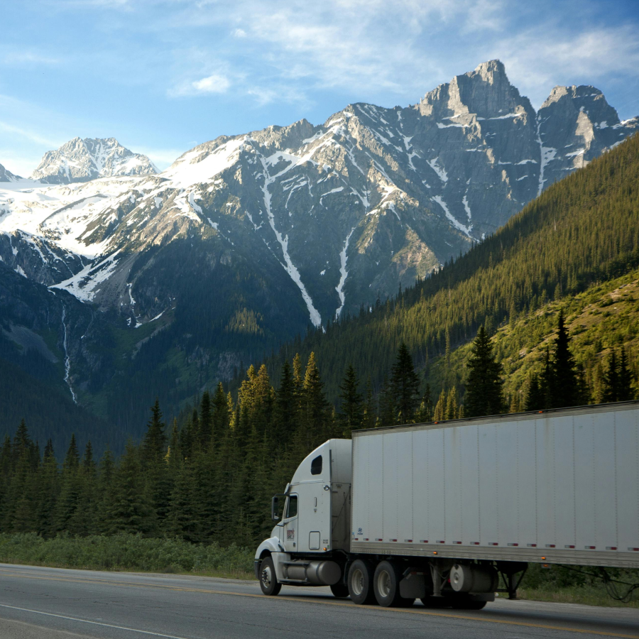 White semi-truck on a mountain road with snow-capped peaks and forests.