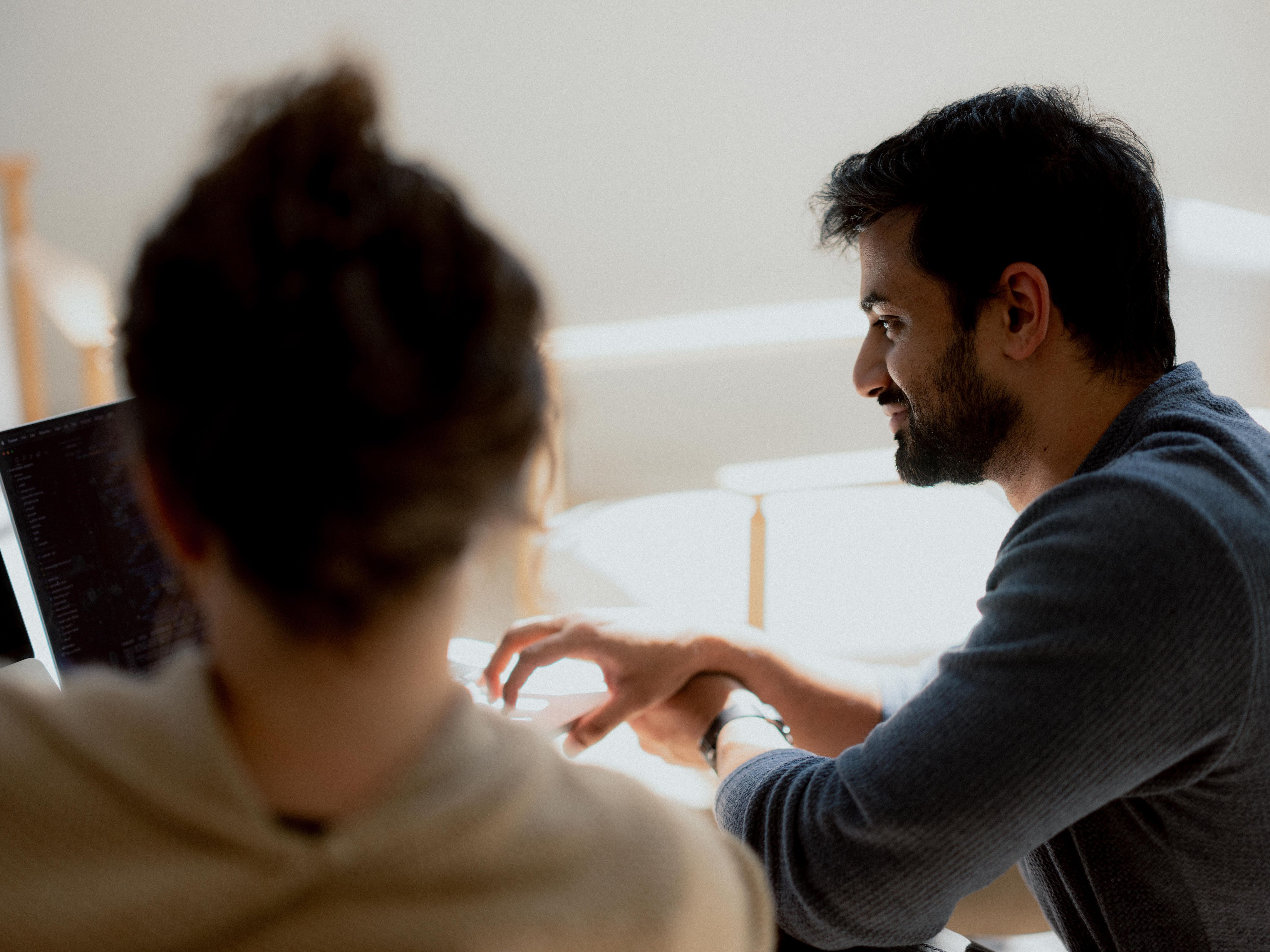 Two people working together at a desk, one using a laptop while the other gestures and smiles during the discussion in a bright, naturally lit room.