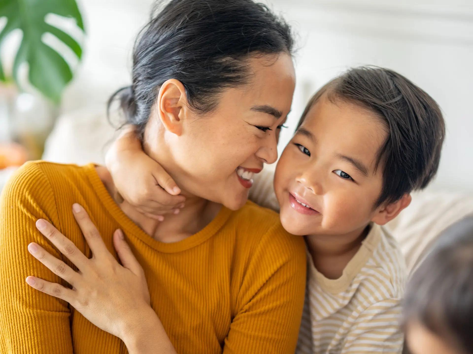 A smiling Asian woman embraces a young boy, who smiles at the camera.