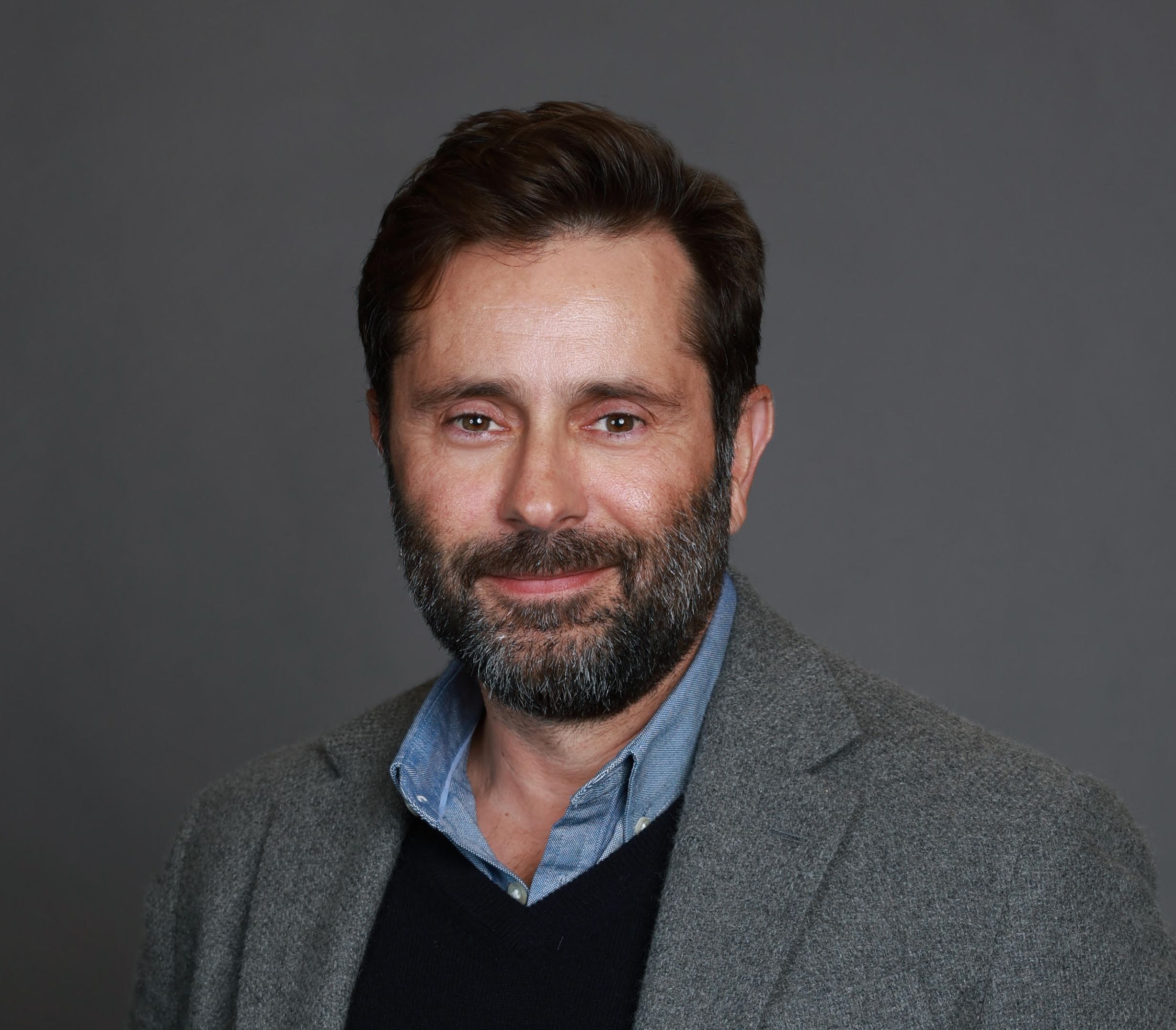 A smiling man with a beard and dark hair, wearing a blue shirt, dark sweater, and grey blazer.